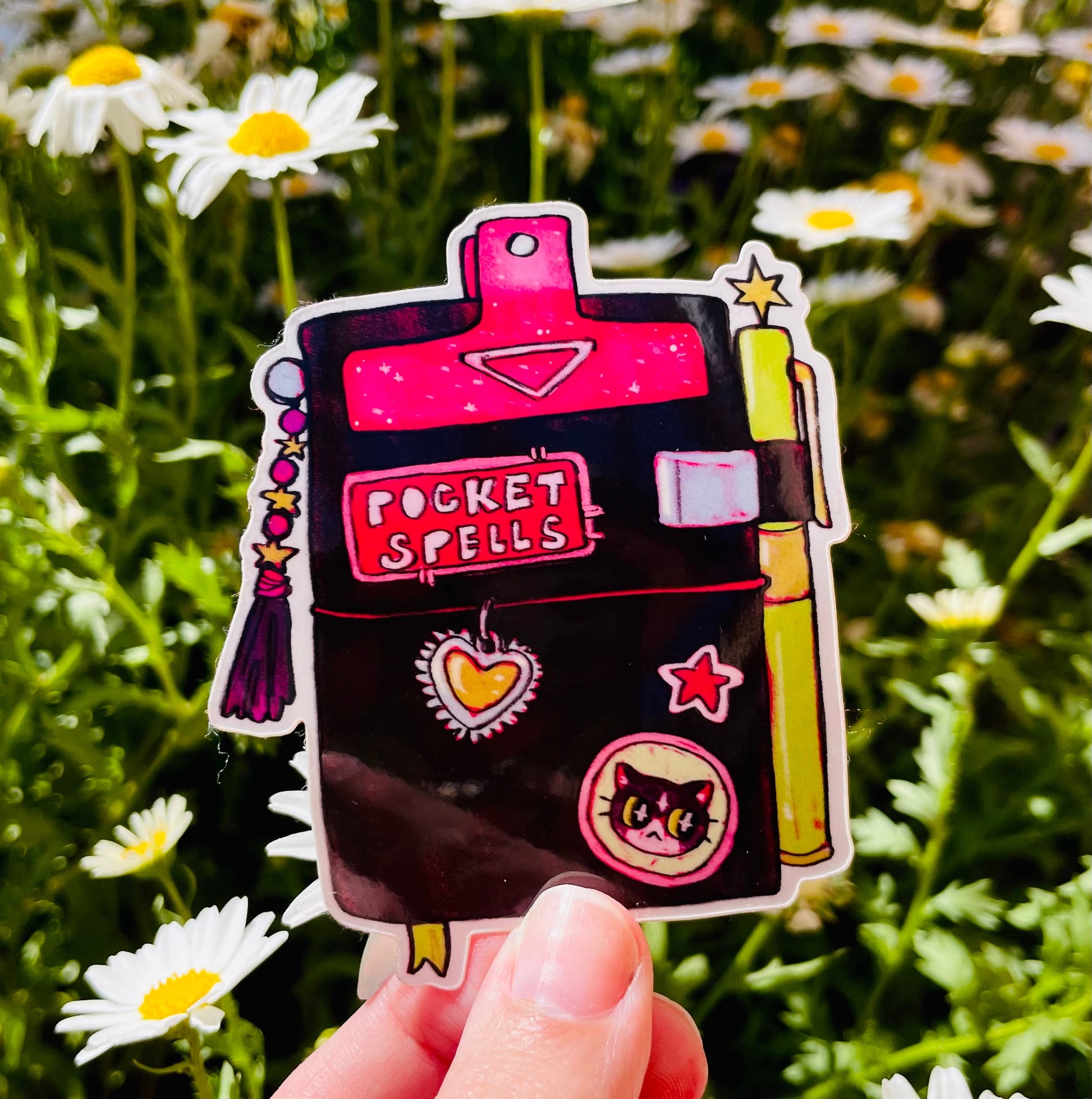 Sticker with 'Pocket Spells' design held in front of a field of daisies