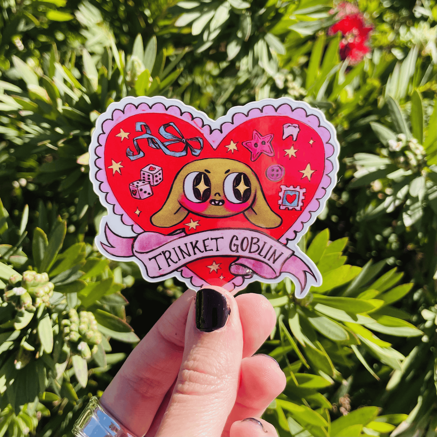 Sticker with cartoon character and 'Trinket Goblin' text held in front of green foliage